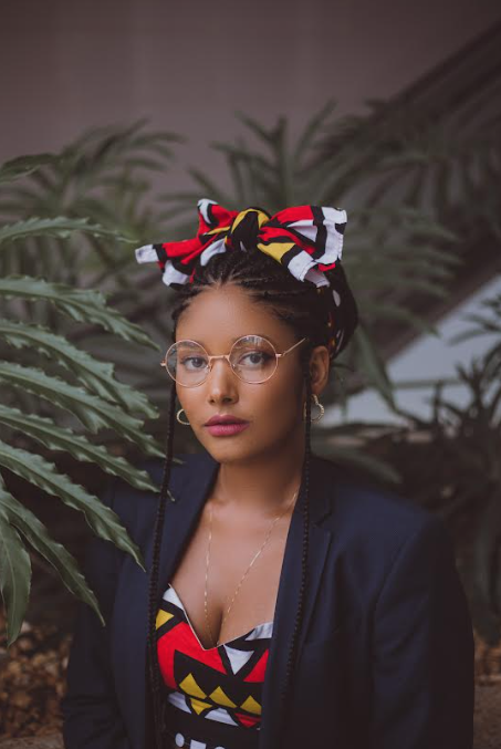 Uma mulher negra, Keilla Vila Flor, está posando em um ambiente interno com plantas ao fundo. Ela usa um lenço colorido em tons de vermelho, amarelo, branco e preto, combinando com sua roupa estampada. Seus óculos redondos e brincos dourados complementam o visual. Ela veste um blazer azul-escuro e mantém uma expressão confiante e séria. Sua luta antirracista está nas redes sociais.