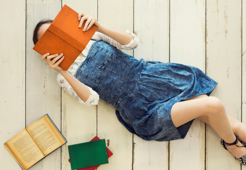 Uma jovem deitada no chão de madeira branca, lendo um livro de capa laranja que cobre parcialmente seu rosto. Ela está vestindo um vestido jeans e uma blusa branca com detalhes sutis. Ao lado dela, há três livros espalhados: um aberto com páginas amareladas e dois fechados, um verde e outro vermelho. A cena transmite uma atmosfera tranquila e descontraída, com um foco na leitura e no prazer da literatura.