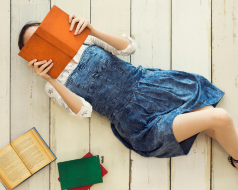 Uma jovem deitada no chão de madeira branca, lendo um livro de capa laranja que cobre parcialmente seu rosto. Ela está vestindo um vestido jeans e uma blusa branca com detalhes sutis. Ao lado dela, há três livros espalhados: um aberto com páginas amareladas e dois fechados, um verde e outro vermelho. A cena transmite uma atmosfera tranquila e descontraída, com um foco na leitura e no prazer da literatura.