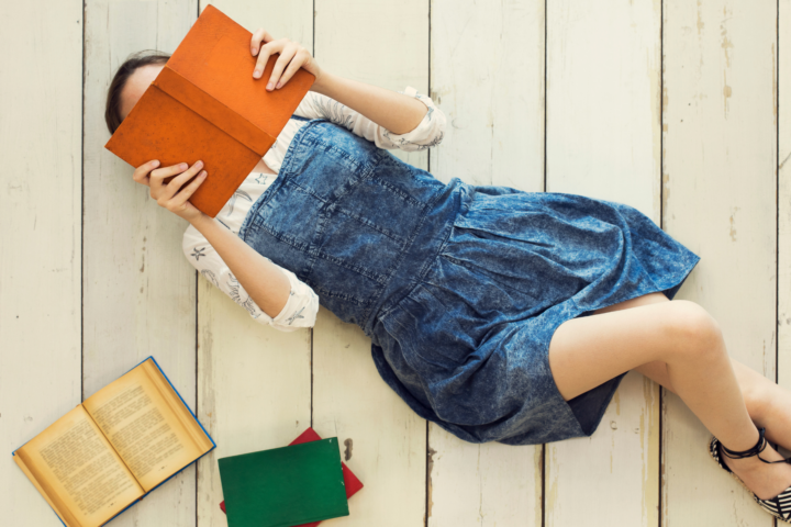 Uma jovem deitada no chão de madeira branca, lendo um livro de capa laranja que cobre parcialmente seu rosto. Ela está vestindo um vestido jeans e uma blusa branca com detalhes sutis. Ao lado dela, há três livros espalhados: um aberto com páginas amareladas e dois fechados, um verde e outro vermelho. A cena transmite uma atmosfera tranquila e descontraída, com um foco na leitura e no prazer da literatura.