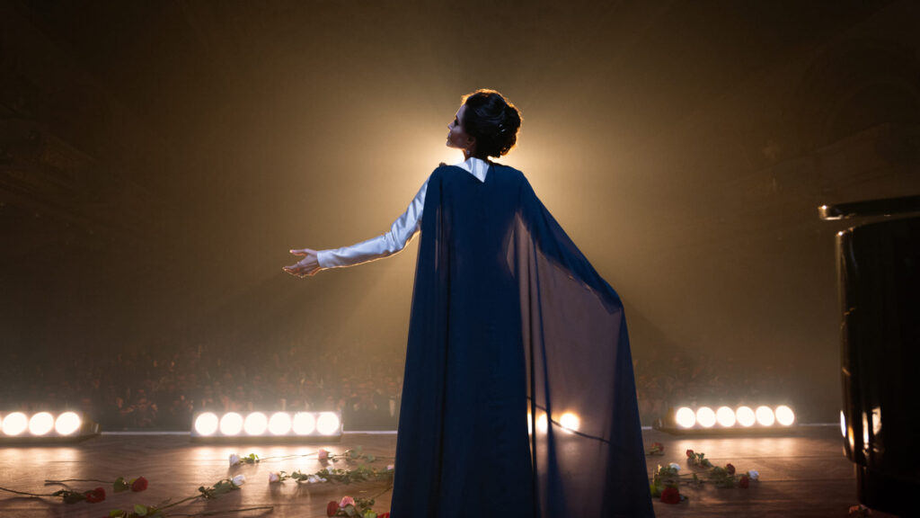 Angelina Julie interpreta Maria Callas em cena do filme. Ela aparece de costas. A luz é escura e há um feixe de luz no palco.