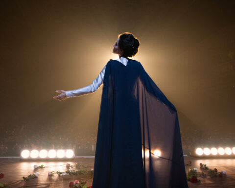 Angelina Julie interpreta Maria Callas em cena do filme. Ela aparece de costas. A luz é escura e há um feixe de luz no palco.