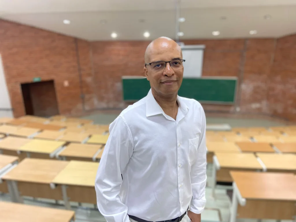 Professor Marcos Aurélio de branco em uma sala de aula. O professor é um homem negro de meia idade.