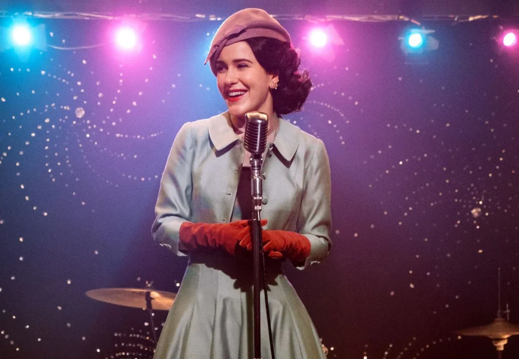 Cena de "A Maravilhosa Mrs. Maisel". Mulher de cerca de 30 anos sorri em um palco iluminado com luzes roxas e azuis.